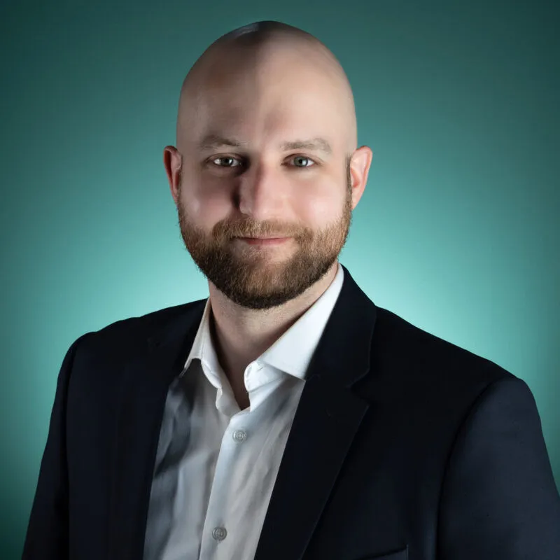 Bald man with a beard smiling in a dark blazer against a teal background