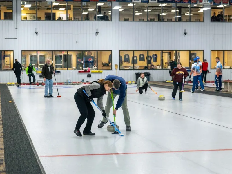 Two curlers sweep the ice at an indoor curling club while other players compete on adjacent lanes