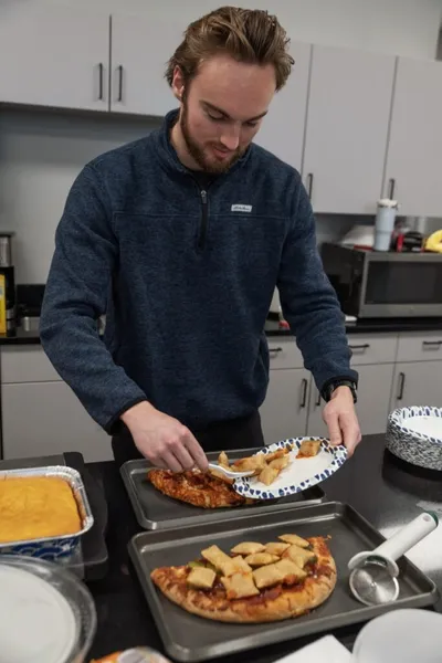 Young man in a navy fleece serving toppings onto a flatbread pizza at a kitchen counter