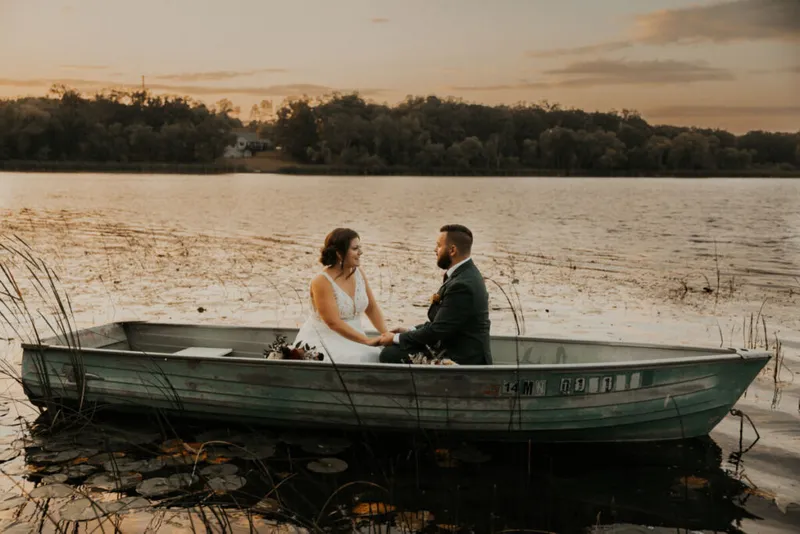 Bride in white gown and groom in dark suit sit facing each other in a small rowboat on a calm lake at sunset