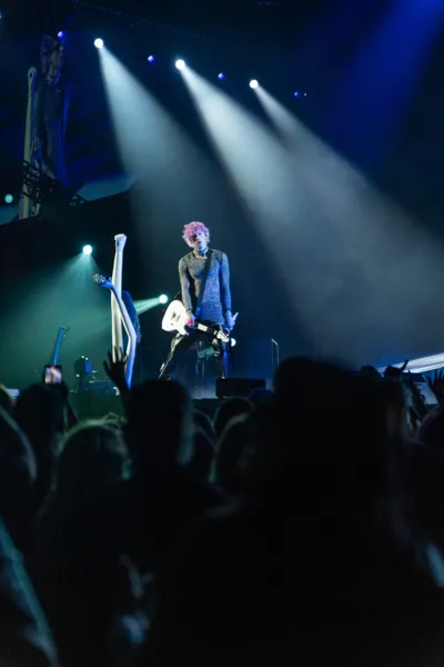 Pink-haired performer holding a white guitar on a lit stage above a crowd