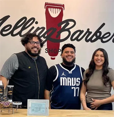 Three smiling people posing behind a counter inside a barber shop