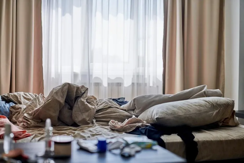 Unmade bed with tangled sheets and pillows beside a cluttered bedside table