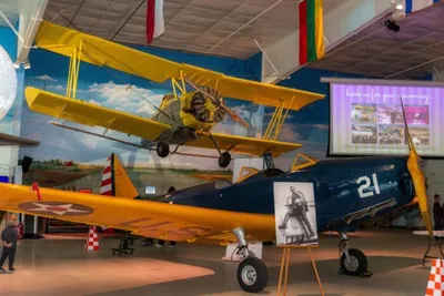 Yellow biplane suspended above a blue Navy aircraft numbered 21 inside an aviation museum