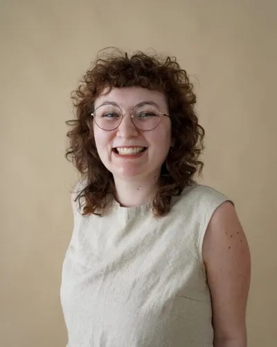Smiling woman with curly auburn hair and round glasses wearing a sleeveless light green top