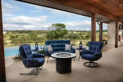 Covered patio with navy cushioned chairs and loveseat around a round fire pit overlooking a pool and rolling landscape