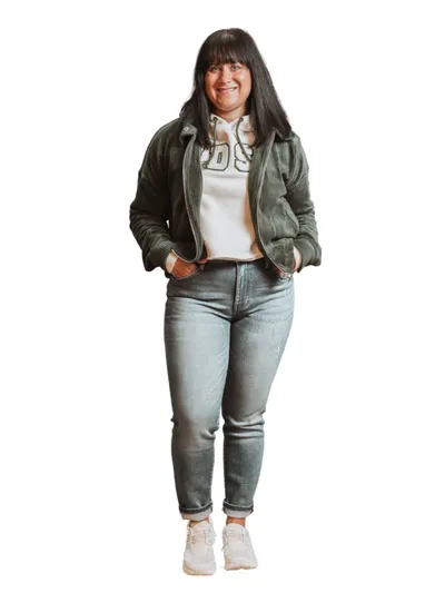 Woman in olive bomber jacket, graphic hoodie, and light-wash jeans smiling on white background