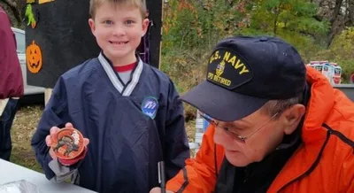 Smiling boy in hockey jersey holds a small decorated pumpkin beside an older man in a U.S. Navy cap at an outdoor fall event