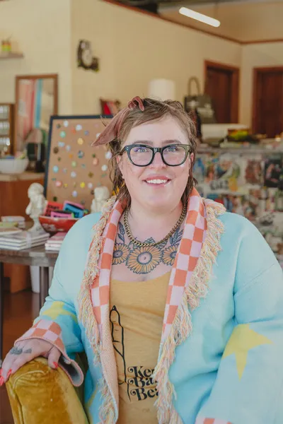 Smiling woman with glasses and neck tattoo wearing a light blue jacket and checkered scarf inside a colorful shop