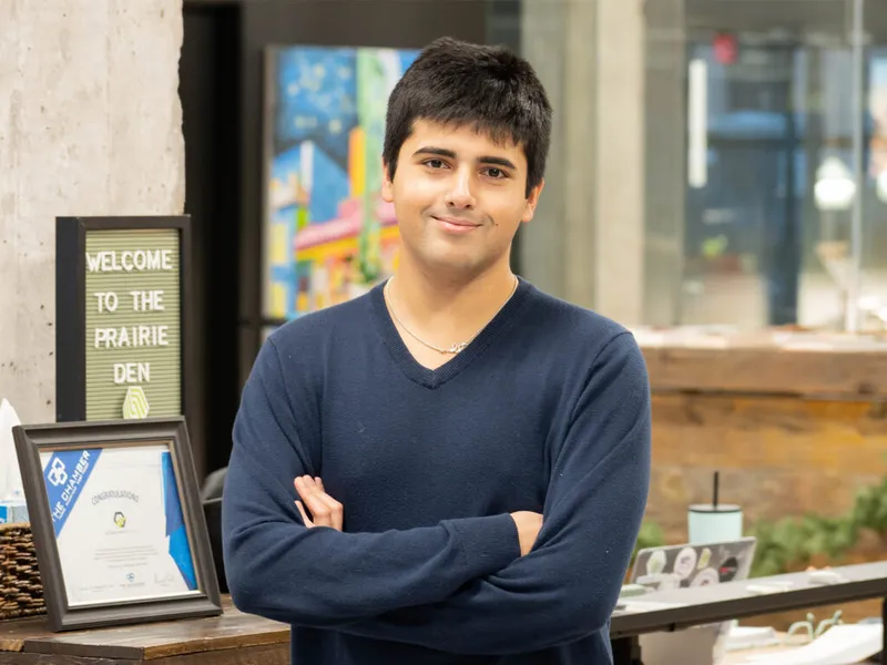 Young man in navy sweater stands arms crossed in front of a 'Welcome to the Prairie Den' sign