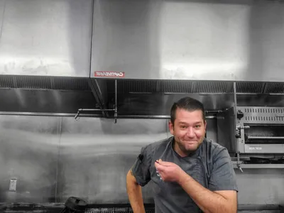 Smiling man in gray chef's shirt posing in a stainless steel commercial kitchen