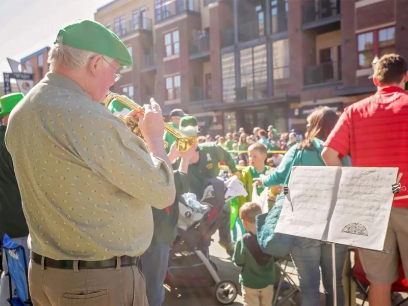 Man in green beret plays trumpet at a sunny outdoor street parade with crowd behind him