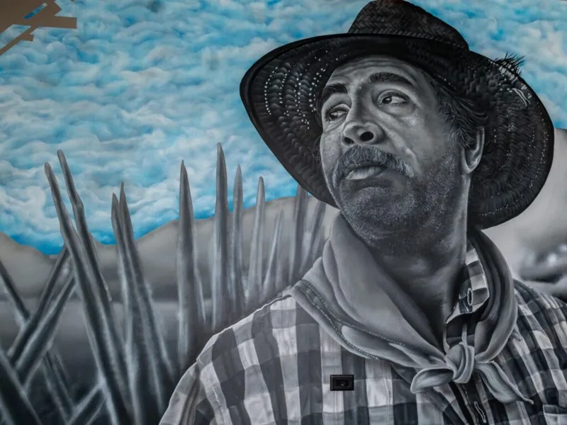 Mural of a man in a straw hat and plaid shirt beside agave spikes under a blue sky