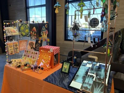 Craft vendor table with orange cloth displaying colorful art prints, pins, and hanging stained glass pieces