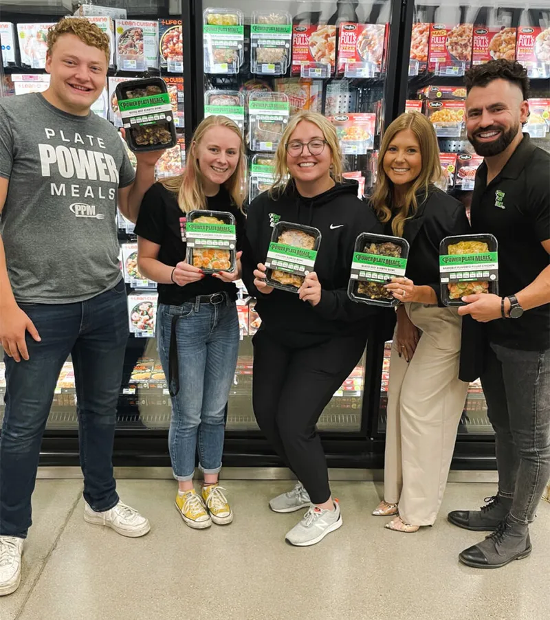 Five smiling people holding Power Plate Meals packaged food in a grocery store freezer aisle