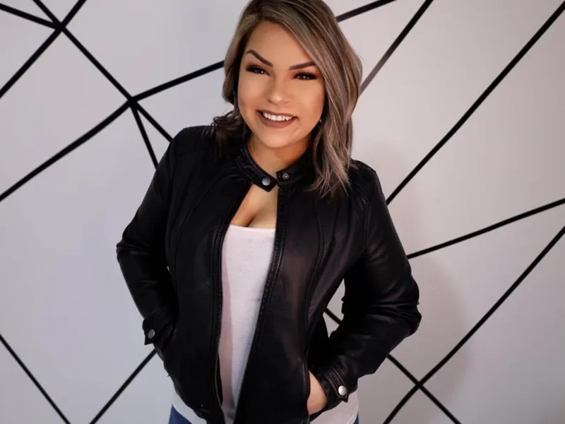 Smiling woman in a black leather jacket and white top posing against a geometric wall