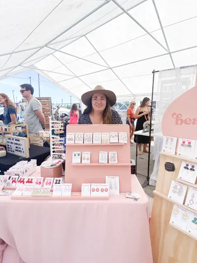 Smiling woman in a wide-brim hat stands behind a pink table displaying handmade earrings at an outdoor market