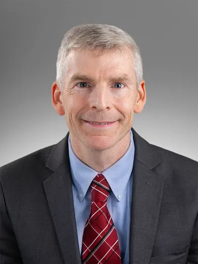 Man with short gray hair in a dark blazer, blue shirt, and red plaid tie