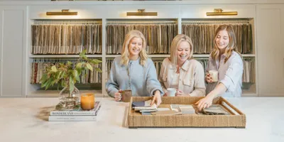 Three women smile around a marble counter holding fabric swatches, with shelving of textile samples behind them