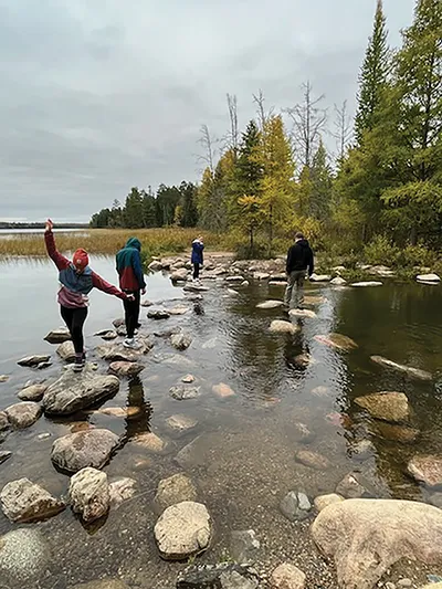 Four people stepping across rocks over a shallow stream lined with evergreens