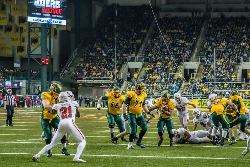NDSU Bison football players in green and gold run a play against red-uniformed opponents in a packed indoor stadium