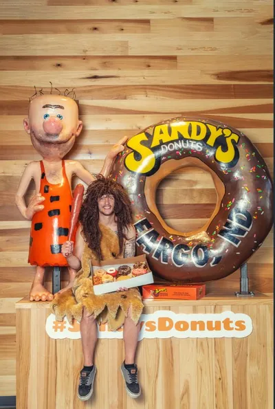Person in caveman costume holds a donut box beside a large Sandy's Donuts mascot statue and giant donut prop