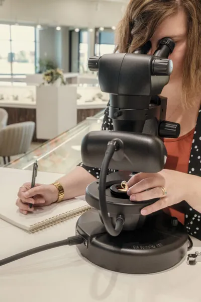Woman examining a gold ring under a gemological microscope while taking notes in a jewelry store
