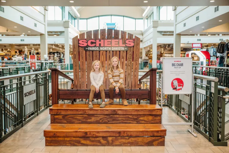 Two young girls sitting on a giant wooden Adirondack chair inside Scheels Fargo