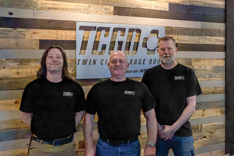 Three men in matching black TCGD shirts standing before a wood-plank wall with a Twin City Garage Door sign