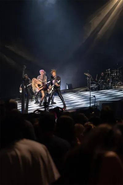 Rock band performing on a checkerboard stage under dramatic spotlights before a packed crowd