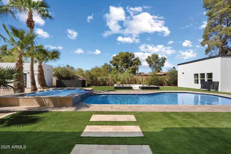 Backyard pool and spa with green lawn, stone path, palm trees, and white stucco exterior under blue sky