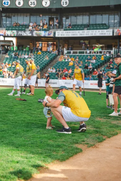 Adult in yellow jersey crouching to help a toddler on a baseball field during a charity event