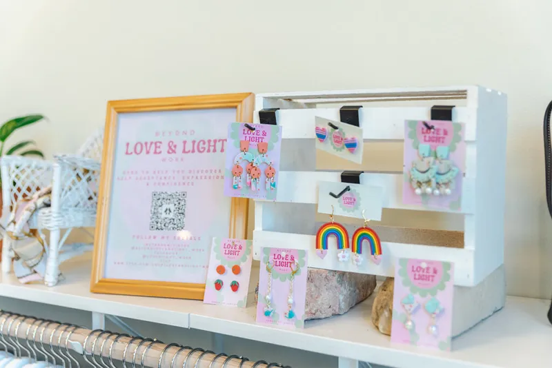 Colorful novelty earrings on pink display cards arranged on a white tiered stand beside a framed Love & Light sign