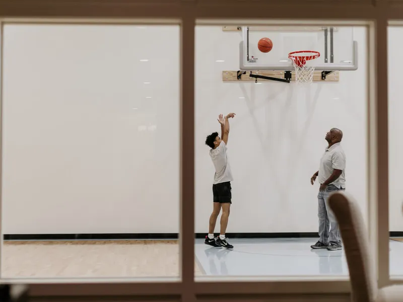 A teen shoots a basketball toward an indoor home court hoop while an adult watches