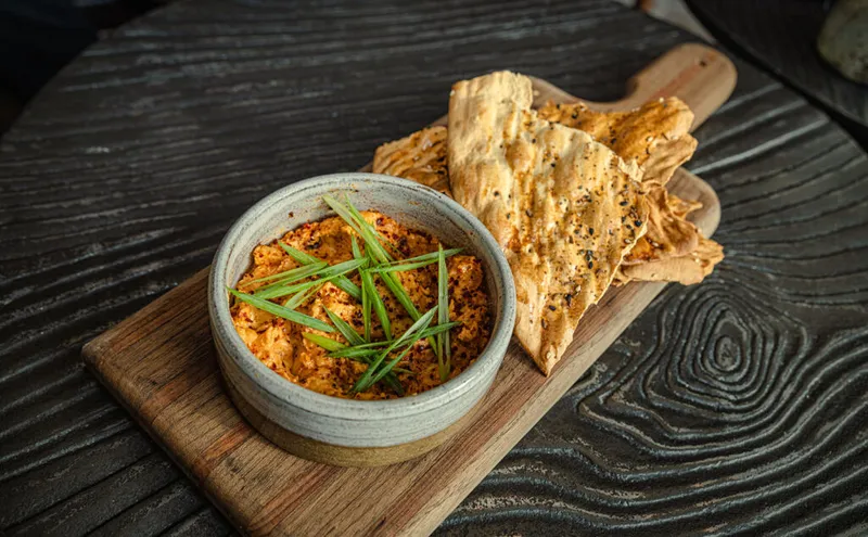 Spiced orange dip topped with green onions in a ceramic bowl, served with seeded flatbread on a wooden board