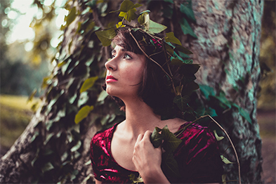 Woman in red velvet dress and ivy leaf crown leaning against a tree