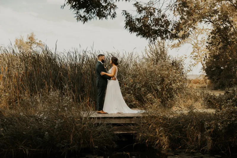 Bride in white gown and groom in dark suit embrace on a wooden dock amid tall reeds
