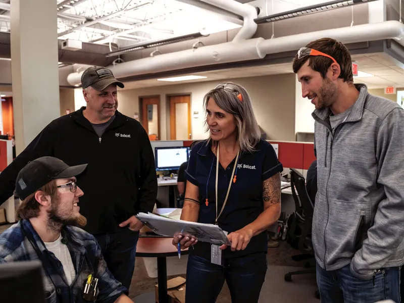Four Bobcat employees in branded uniforms gathered around documents in an open office