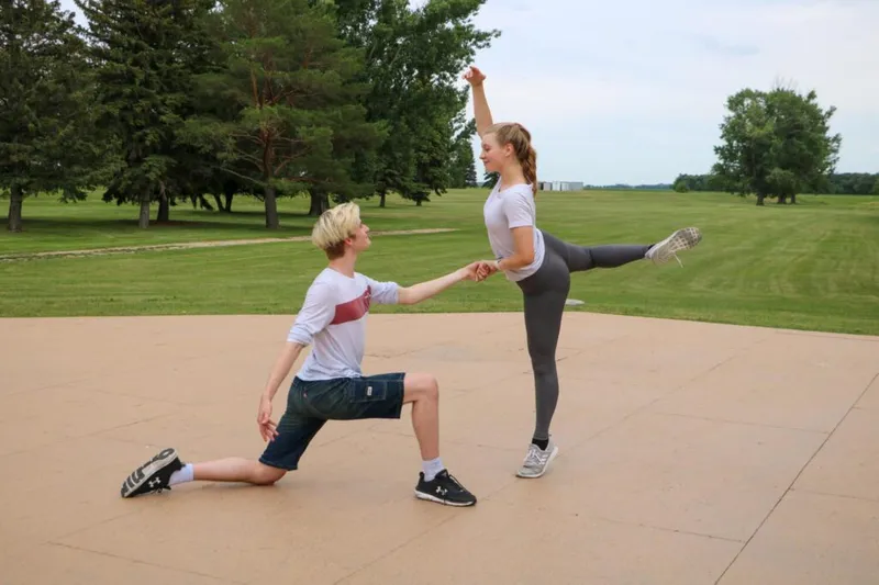Two young dancers hold hands outdoors, one kneeling and one balancing on one leg with arm raised