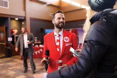 Smiling bearded man in a red blazer speaks into a microphone at an indoor community event