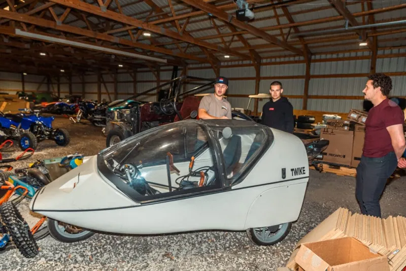 Three men examine a white Twike three-wheeled vehicle inside a large barn workshop