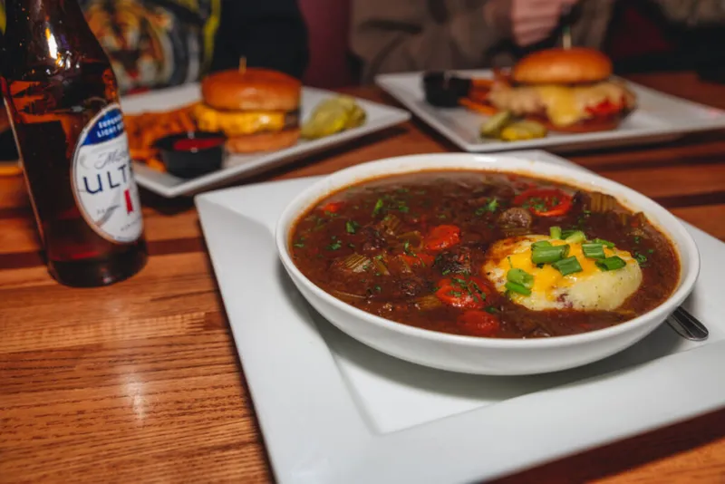 Bowl of rich beef stew topped with mashed potatoes and scallions, with burgers and a beer bottle in the background
