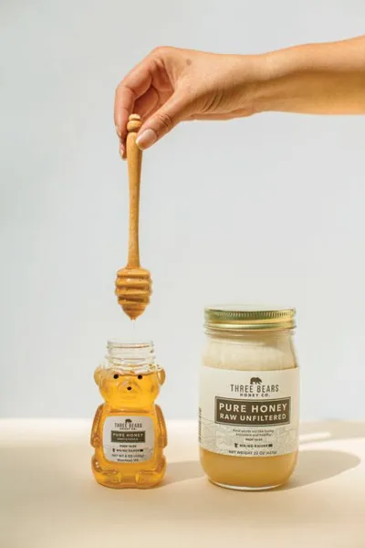 Hand holding a honey dipper over a bear-shaped jar beside a larger Three Bears Pure Honey jar