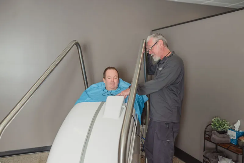 Man in blue robe reclining in a hyperbaric chamber assisted by a staff member