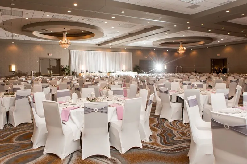 Large ballroom set for a wedding reception with white chair covers, pink and gray accents, and chandelier lighting