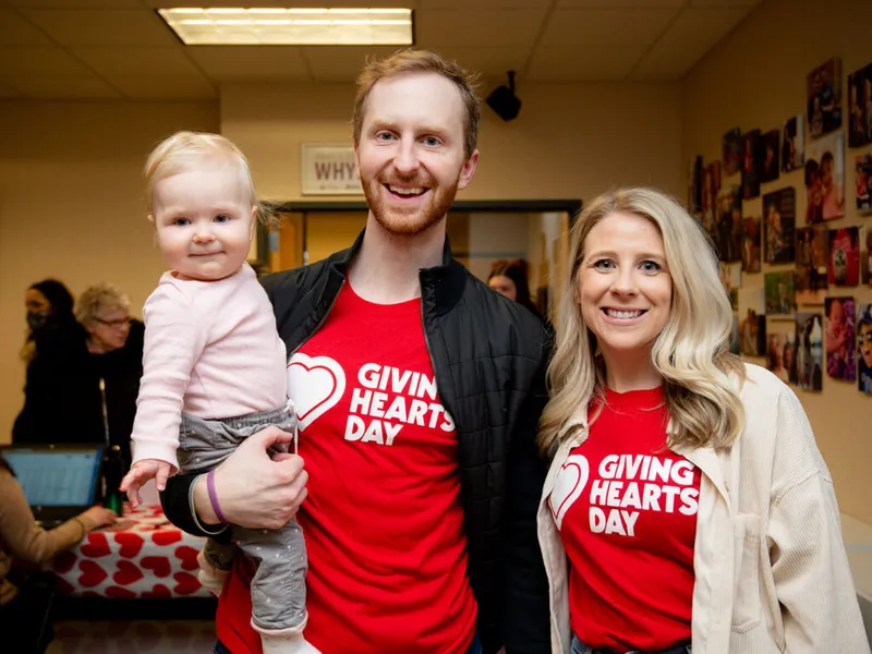 Man and woman in red Giving Hearts Day shirts smiling, man holding a baby