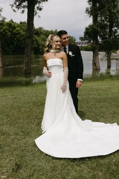 Bride in strapless white gown with train and groom in black suit pose by a river