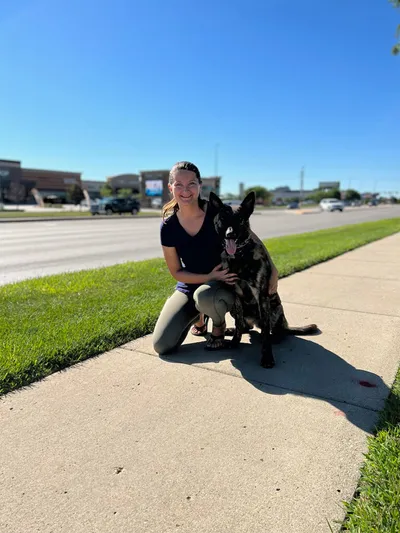 Smiling woman kneeling on a sidewalk beside a large black dog on a sunny day