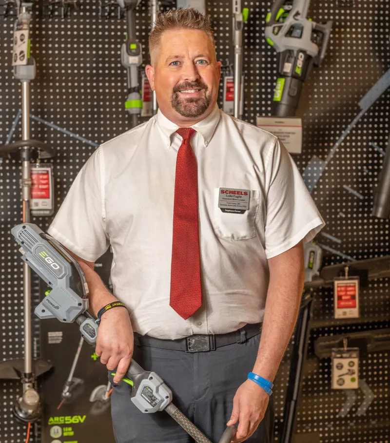 Smiling Scheels employee in white shirt and red tie holding an EGO cordless trimmer in a store display