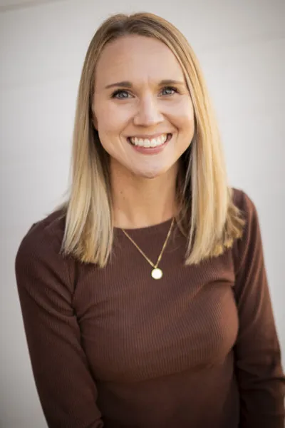 Smiling woman with blonde hair wearing a brown ribbed top and gold pendant necklace